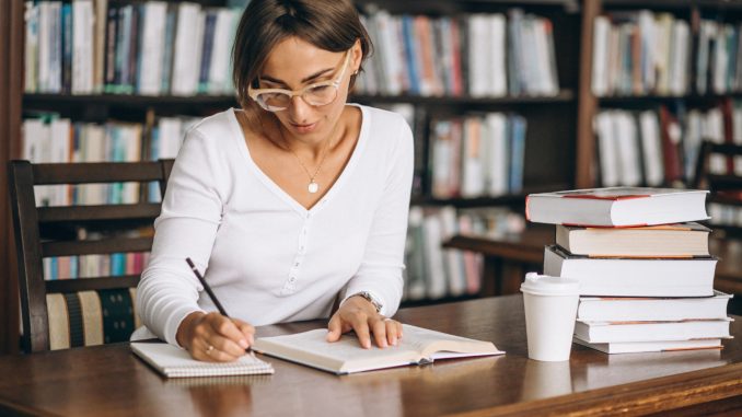 Kobieta robiąca notatki przy biurku w bibliotece, w tle regały z książkami